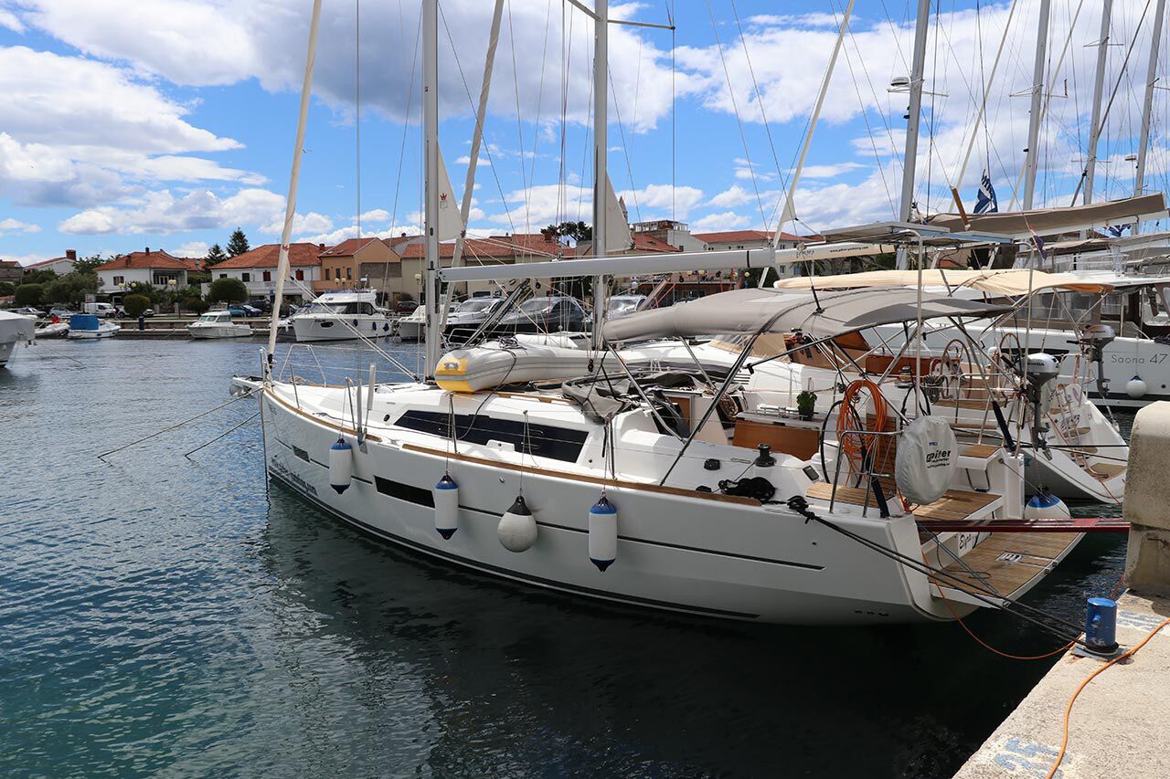 A marina with several sailboats docked, a calm water surface, and buildings in the background.