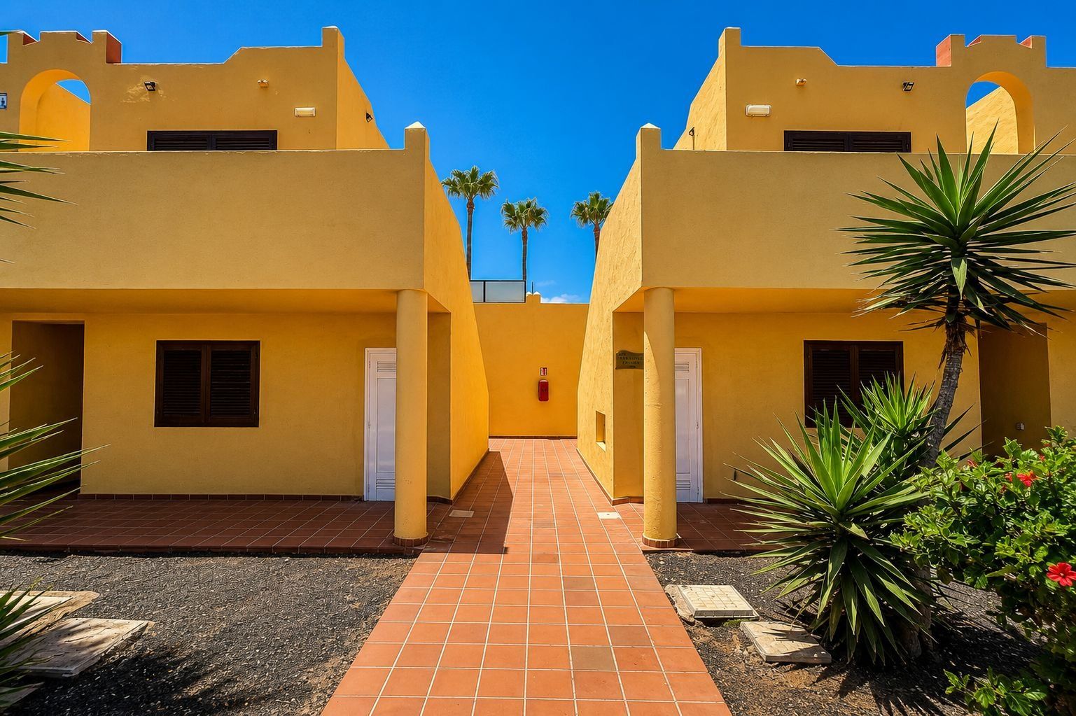 A courtyard with yellow buildings, a tiled walkway, and some greenery.