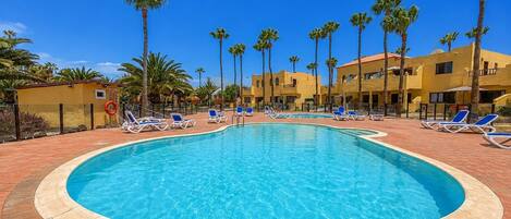 A swimming pool with sun loungers and palm trees, surrounded by yellow buildings.