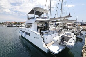 Eine weiße Yacht namens 'MY 37' liegt in einer Marina festgemacht, zusammen mit anderen Booten, mit einer überdachten Deckfläche und Blick auf Gebäude im Hintergrund.