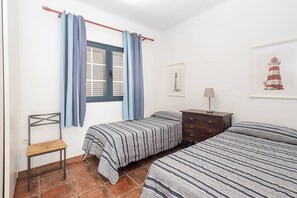 Un dormitori amb dos llits, una cadira, una petita arca i una finestra amb cortines, i una paret amb un quadre emmarcat.