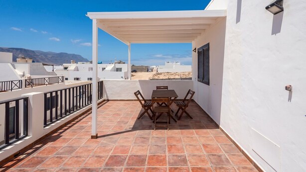 Una terraza en la azotea con una mesa y sillas, paredes blancas, y vistas al mar y a las montañas.