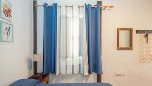 Un dormitorio con cama, mesita de noche, lámpara, ventana con cortinas, espejo adosado a la pared y obras de arte enmarcadas.