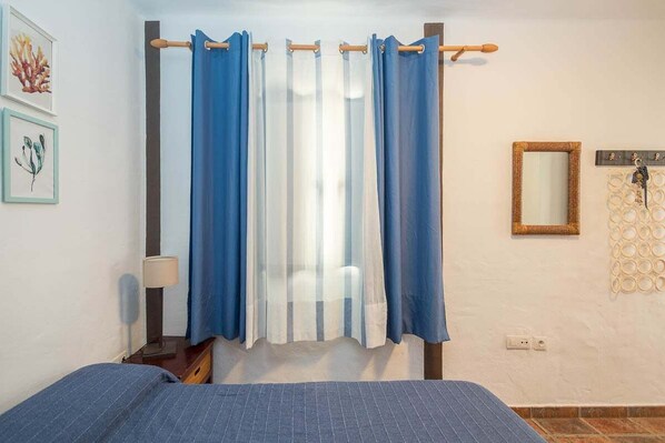 Un dormitori amb un llit, una tauleta de nit, una làmpada, una finestra amb cortines, un mirall fixat a la paret i obra d'art emmarcada.