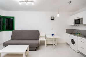 A compact living space with a grey sofa, a small white table, a washing machine, and a microwave in the kitchen area. - GuestReady - Cosy Retreat with Balcony near Beach (El Cotillo)