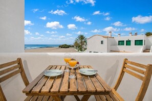 A wooden table with two plates and glasses of orange juice, overlooking a beach and white buildings with green shutters. - GuestReady - Cosy Retreat with Balcony near Beach (El Cotillo)