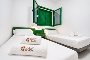 A hotel room with a bed, two pillows, a green-framed window, and a nightstand with a lamp. - GuestReady - Cosy Retreat with Balcony near Beach (El Cotillo)
