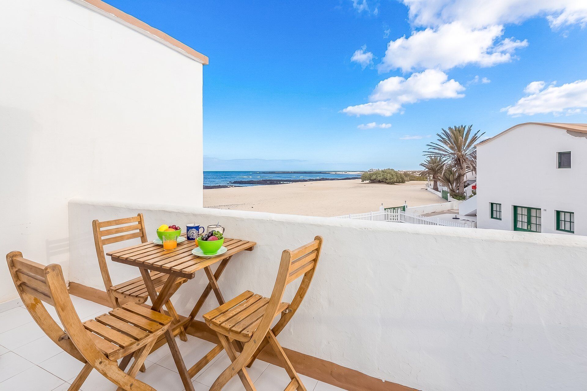 Ein Balkon mit Tisch und Stühlen, Blick auf den Strand, und ein weißes Gebäude mit grünen Fenstern.