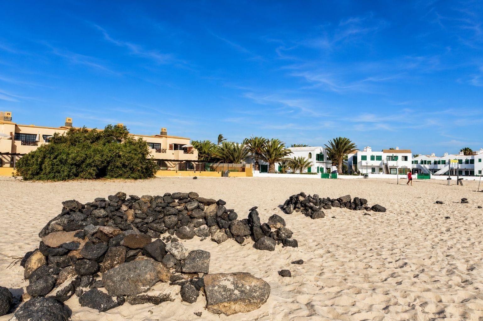 A sandy beach with a pile of rocks in the foreground, buildings with balconies in the background, and a clear blue sky.