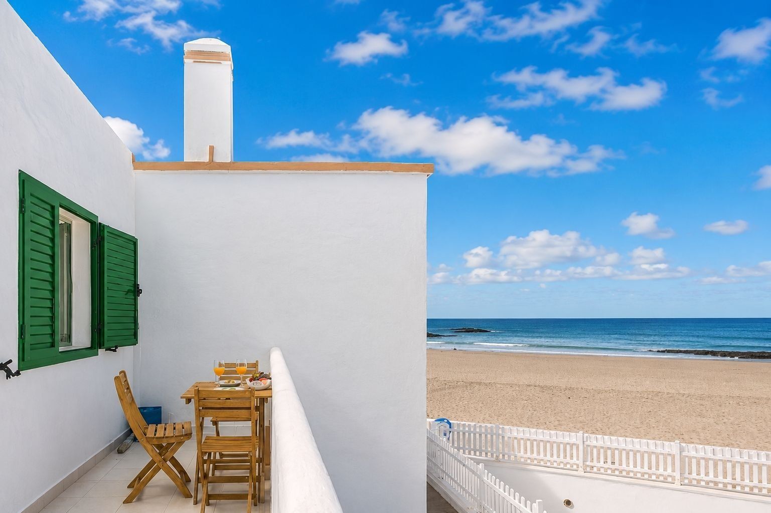 A balcony with a table and chairs, overlooking a beach with a white fence and a clear sky.