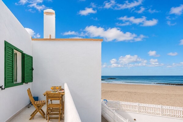A balcony with a table and chairs, overlooking a beach with a white fence and a clear sky. - GuestReady - Cosy Retreat with Balcony near Beach (El Cotillo)