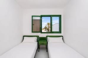 A room with two beds, a small table, and a window with a view of palm trees and a building. - GuestReady - Cosy Retreat with Balcony near Beach (El Cotillo)