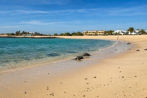 A beach with clear water, a sandy shore, and buildings in the background. - GuestReady - Cosy Retreat with Balcony near Beach (El Cotillo)