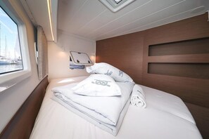 Cama bem arrumada com lençóis brancos e almofadas decorativas, janela com vista para barcos e cabeceira de madeira.