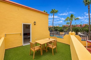 A yellow building with a patio area featuring a table and chairs, a pool, and palm trees in the background. - GuestReady - Bright Retreat with Outdoor Pool (Corralejo)