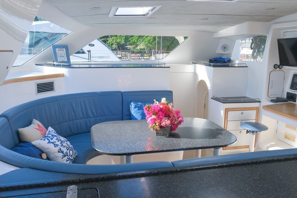 Interior de barco com um sofá azul, uma mesa pequena com um vaso de flores, uma cozinha e uma televisão.