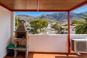 A balcony with a barbecue grill, a trash bin, and a view of a town with mountains in the background. - GuestReady - Gadi House Rural Retreat (Tasarte)