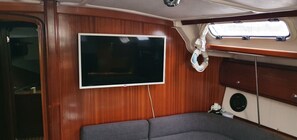A cabin with a flat-screen TV mounted on a wooden wall, a gray sofa, and a window with a view of the sky.