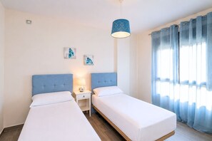 A hotel room with two single beds, a nightstand with a lamp, a wall-mounted light fixture, and a window with curtains. - GuestReady - Pueblo Majorero Sunny Stay and Pool (Antigua)