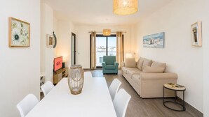 A modern living room with a white dining table, a sofa, and a chair. There is a wicker vase on the table, a lamp, and a painting on the wall. - GuestReady - Pueblo Majorero Sunny Stay and Pool (Antigua)