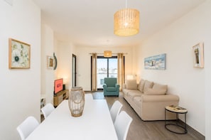 Ein modernes Wohnzimmer mit einem weißen Esstisch, einer Couch und einem Sessel. Auf dem Tisch steht eine Korbvase, eine Lampe und an der Wand hängt ein Gemälde.