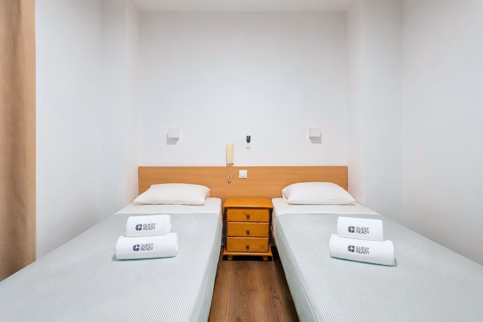 A hotel room with two single beds, a wooden nightstand, a wall-mounted phone, and a 'GUEST READY' sign on each bed.
