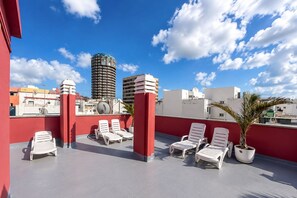 Eine Dachterrasse mit weißen Liegestühlen, roten Säulen und Blick auf Stadtgebäude.