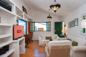 A furnished living room with a television, a sofa, a chair, and a table. There are shelves with books and a lamp on a side table. - GuestReady - Casa Carmen Terrace Sea View (Agaete)