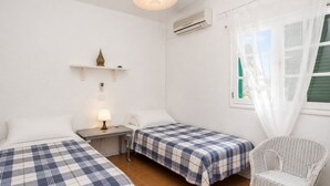 Ein Zimmer mit zwei Betten, einem Sessel, einem Nachttisch mit Lampe, einem Regal mit Dekoration und einem Fenster mit Vorhängen.