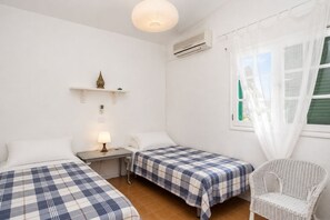 Ein Zimmer mit zwei Betten, einem Sessel, einem Nachttisch mit Lampe, einem Regal mit Dekoration und einem Fenster mit Vorhängen.