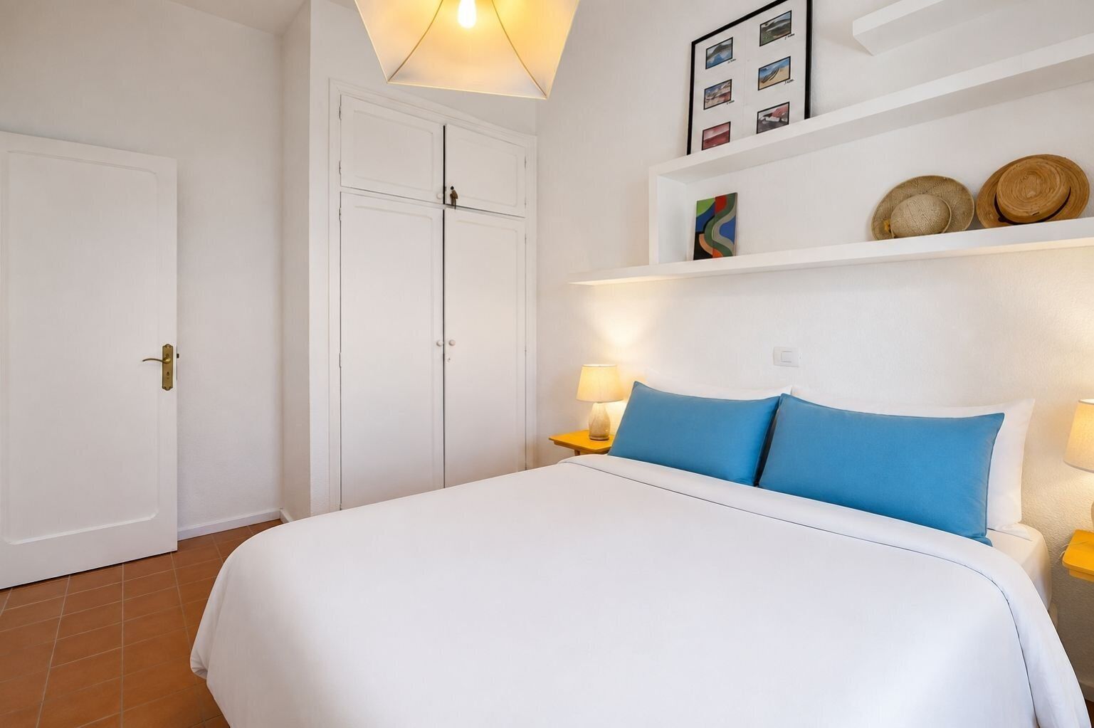 Ett hotellrum med en säng, en garderob, en hylla med hattar och en vägg med inramade bilder.
