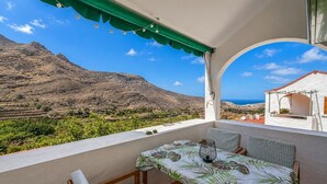 Ein Balkon mit Tisch und Stühlen, der auf eine gebirgige Landschaft und das Meer blickt.