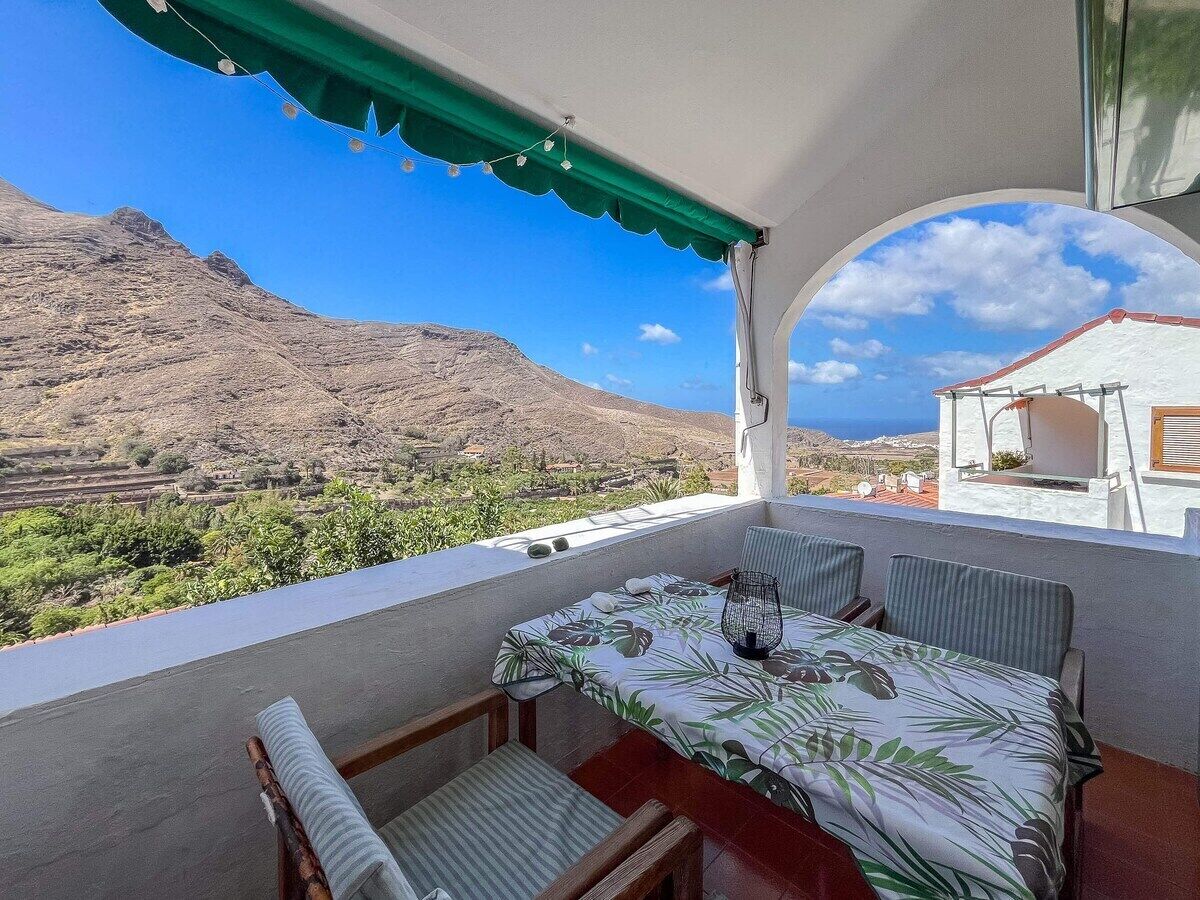 A balcony with a table and chairs, a view of a mountainous landscape, and a building in the background.