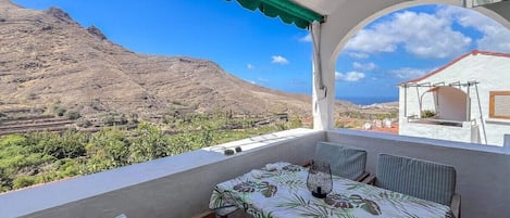 A balcony with a table and chairs, a view of a mountainous landscape, and a building in the background.