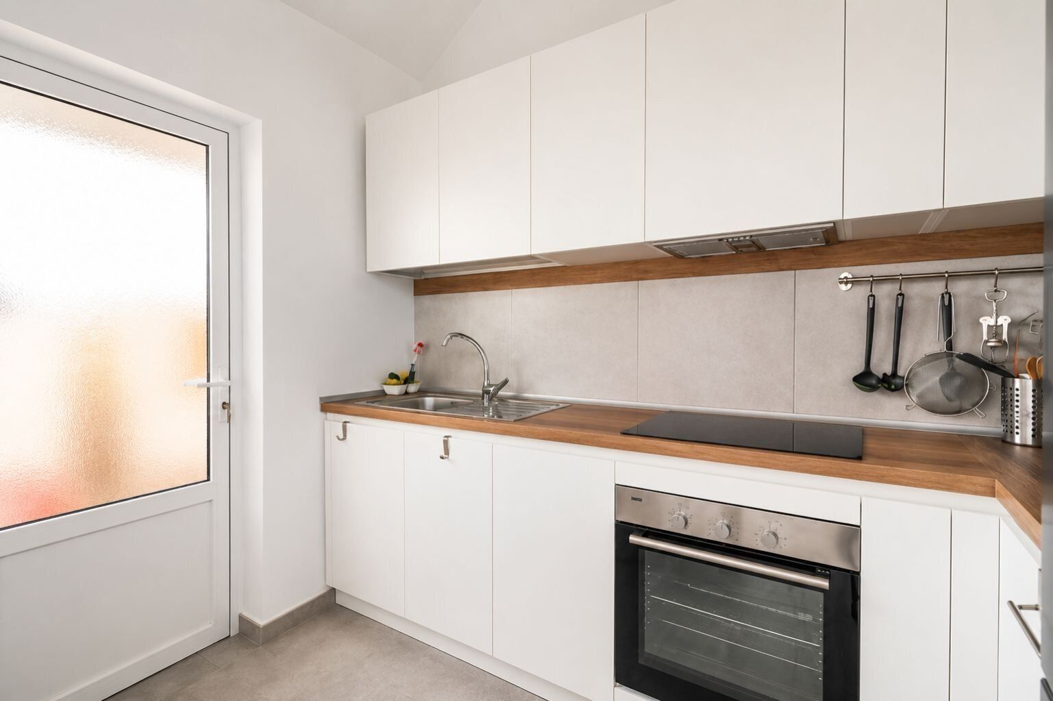 Cocina moderna con armarios blancos, encimera de madera, pileta de acero inoxidable y horno integrado.