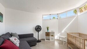A modern living room with a grey sofa, a standing fan, a white baby changing station, and a view of the sky through a window. - GuestReady - Melocotones Family Bungalow (San Bartolomé de Tirajana)