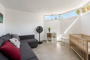 Una sala de estar moderna con un sofá gris, un ventilador de pie, un cambiador para bebés blanco y vistas al cielo a través de una ventana.