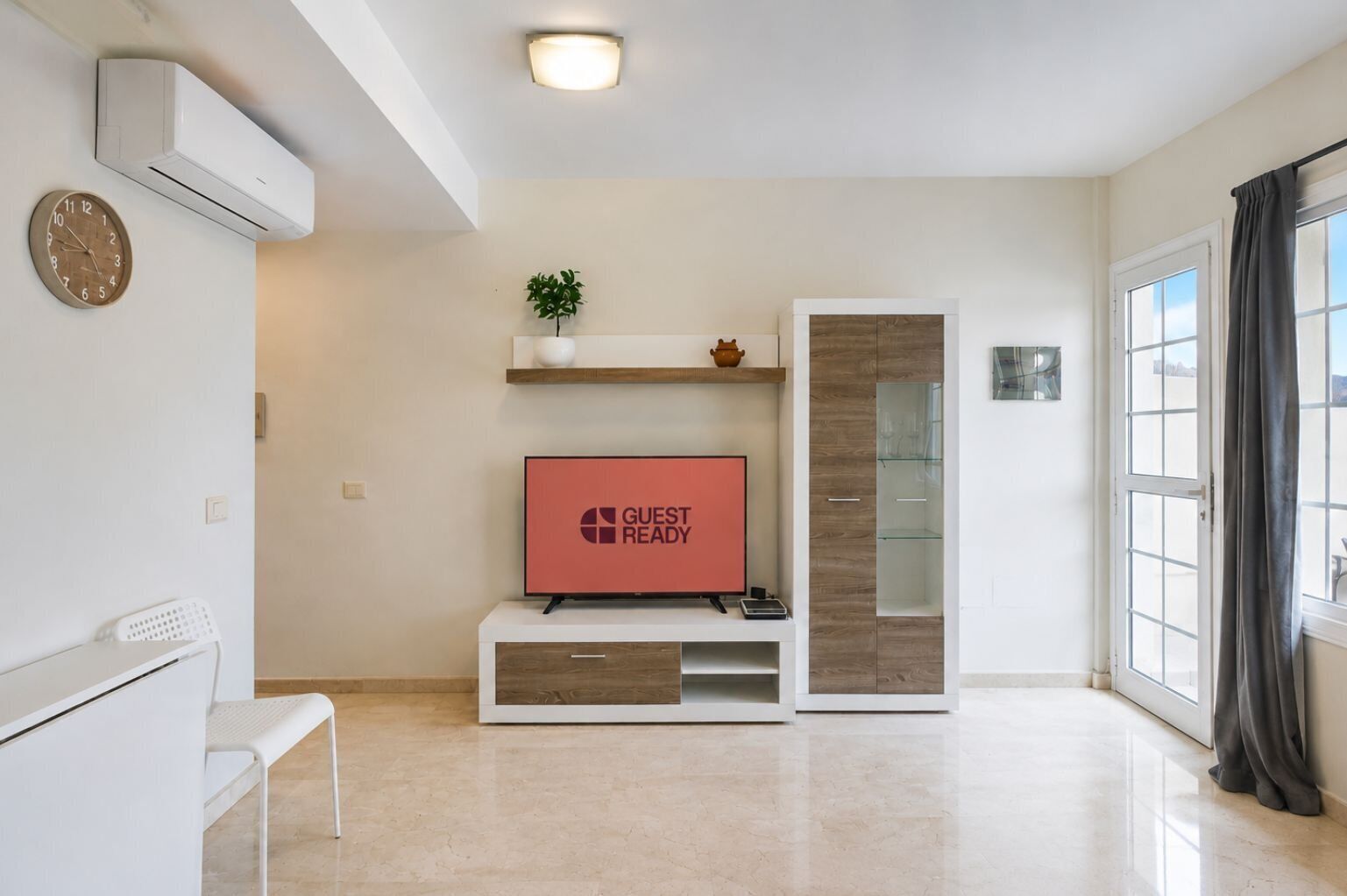 Uma sala de estar moderna com televisão de tela plana, um banco branco, uma prateleira com objetos decorativos e um armário de vidro.