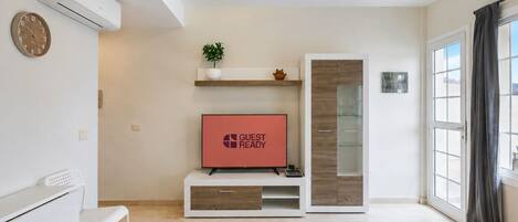 Una sala moderna con un televisor de pantalla plana, un banco blanco, una estantería con adornos y un aparador de vidrio.
