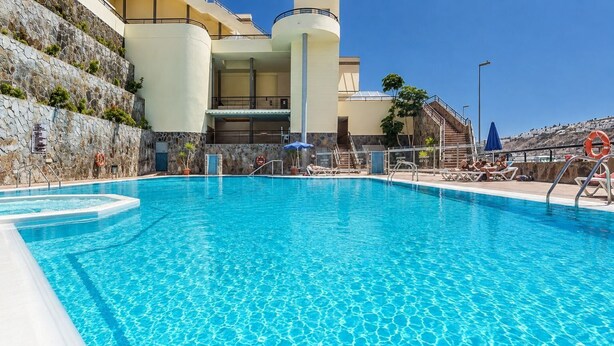 A swimming pool with clear blue water, surrounded by lounge chairs and umbrellas, with a building featuring a stone facade and a balcony in the background. - GuestReady - Puerto Rico Bungalow with Pool (Puerto Rico)