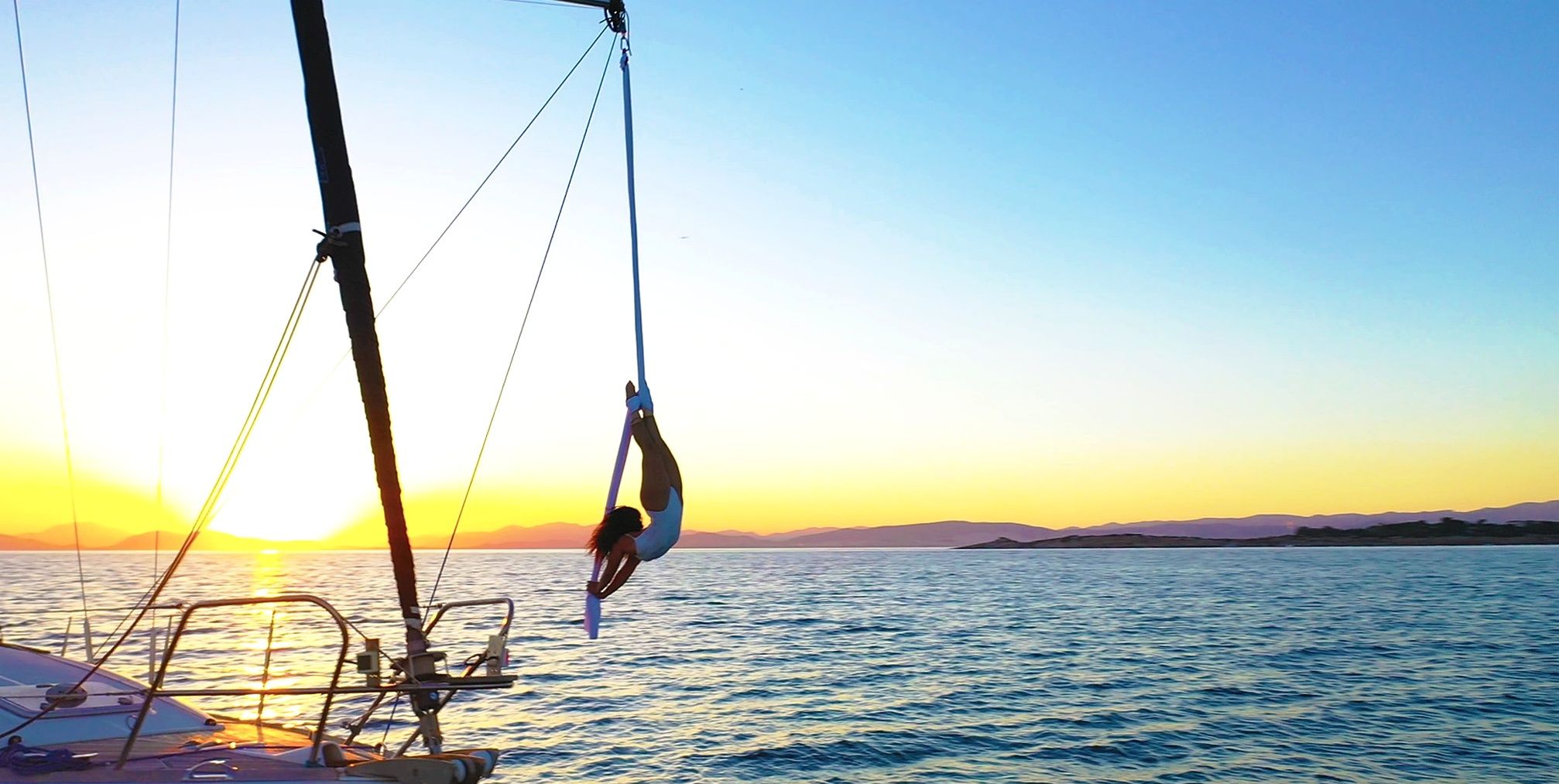 A person performing aerial yoga on a sailboat with a sunset in the background.