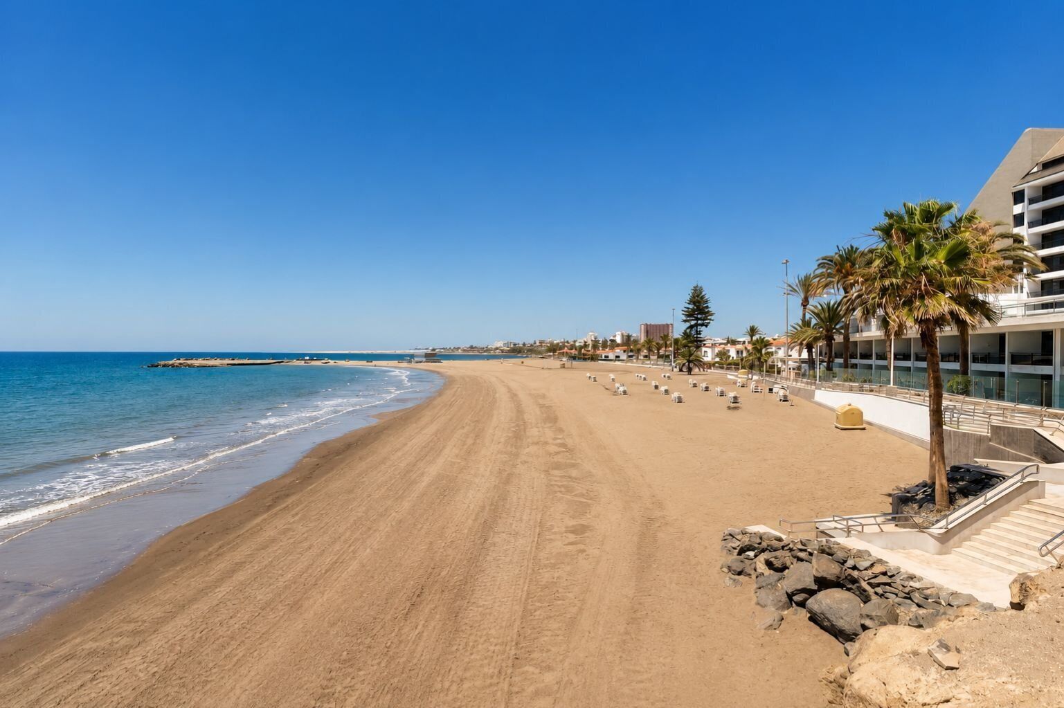 A beach with a clear sky, a sandy shore, and a building with a balcony overlooking the beach.