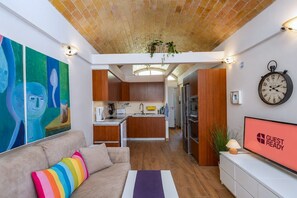 A modern living room with a sofa, a colorful rug, a kitchen area with wooden cabinets, a dining table, and a clock on the wall. - GuestReady - San Agustin Sunny Balcony (San Bartolomé de Tirajana)