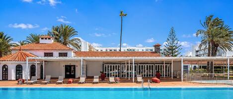 A hotel with a swimming pool, sun loungers, and palm trees.
