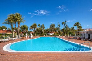 Ein klares, blaues Schwimmbecken, umgeben von Liegestühlen und Palmen, mit einer gefliesten Terrasse und blauem Himmel.