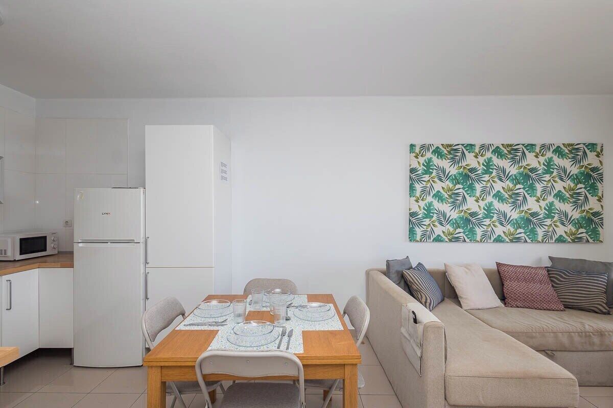 En kompakt bolig med tekøkken, spisebord til fire personer og en sofa med puder.