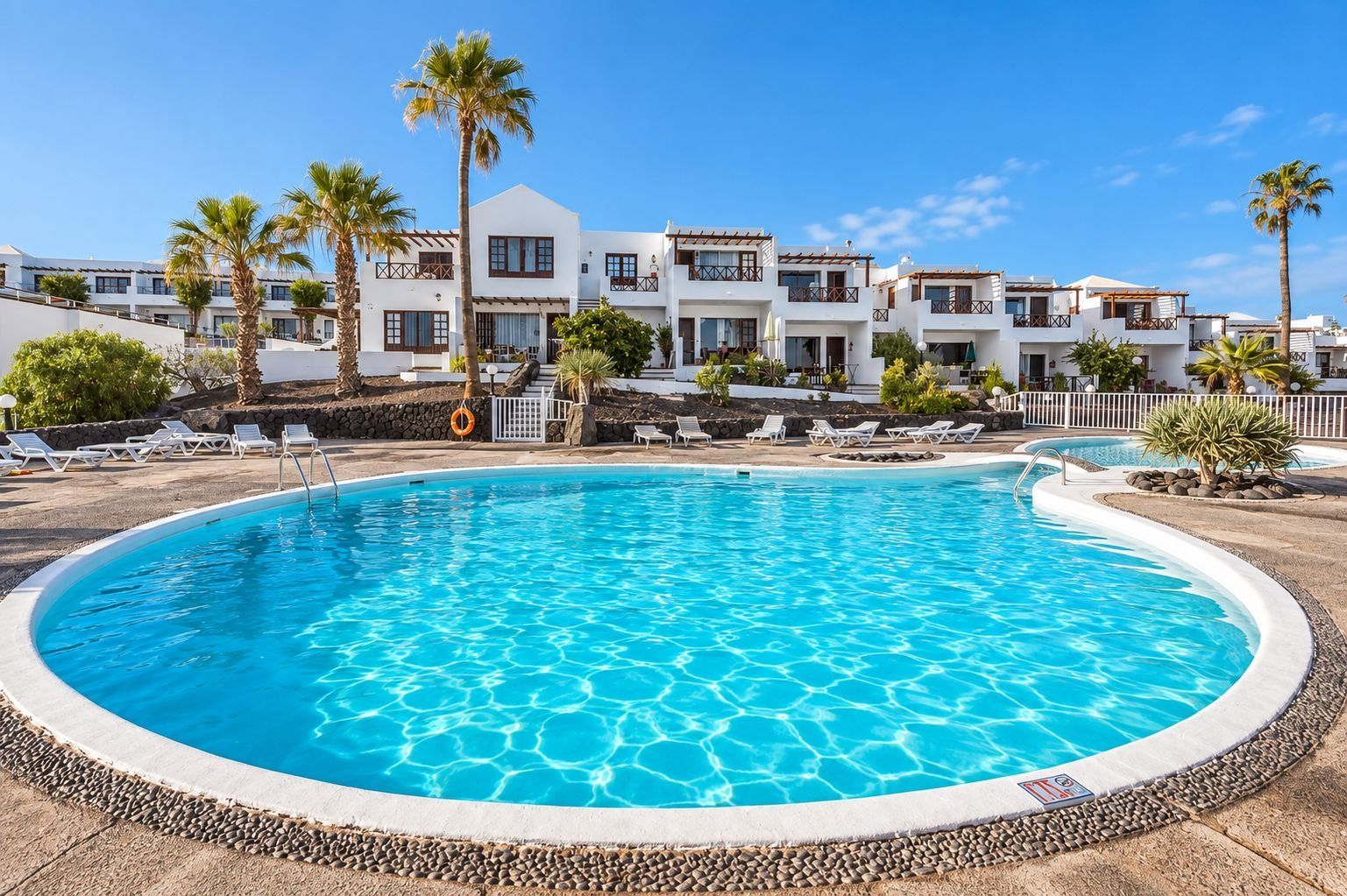 A swimming pool surrounded by lounge chairs and palm trees, with a building in the background.