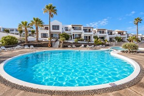 A swimming pool surrounded by lounge chairs and palm trees, with a building in the background. - GuestReady - Las Tabaibas Sea View Retreat (Puerto del Carmen)