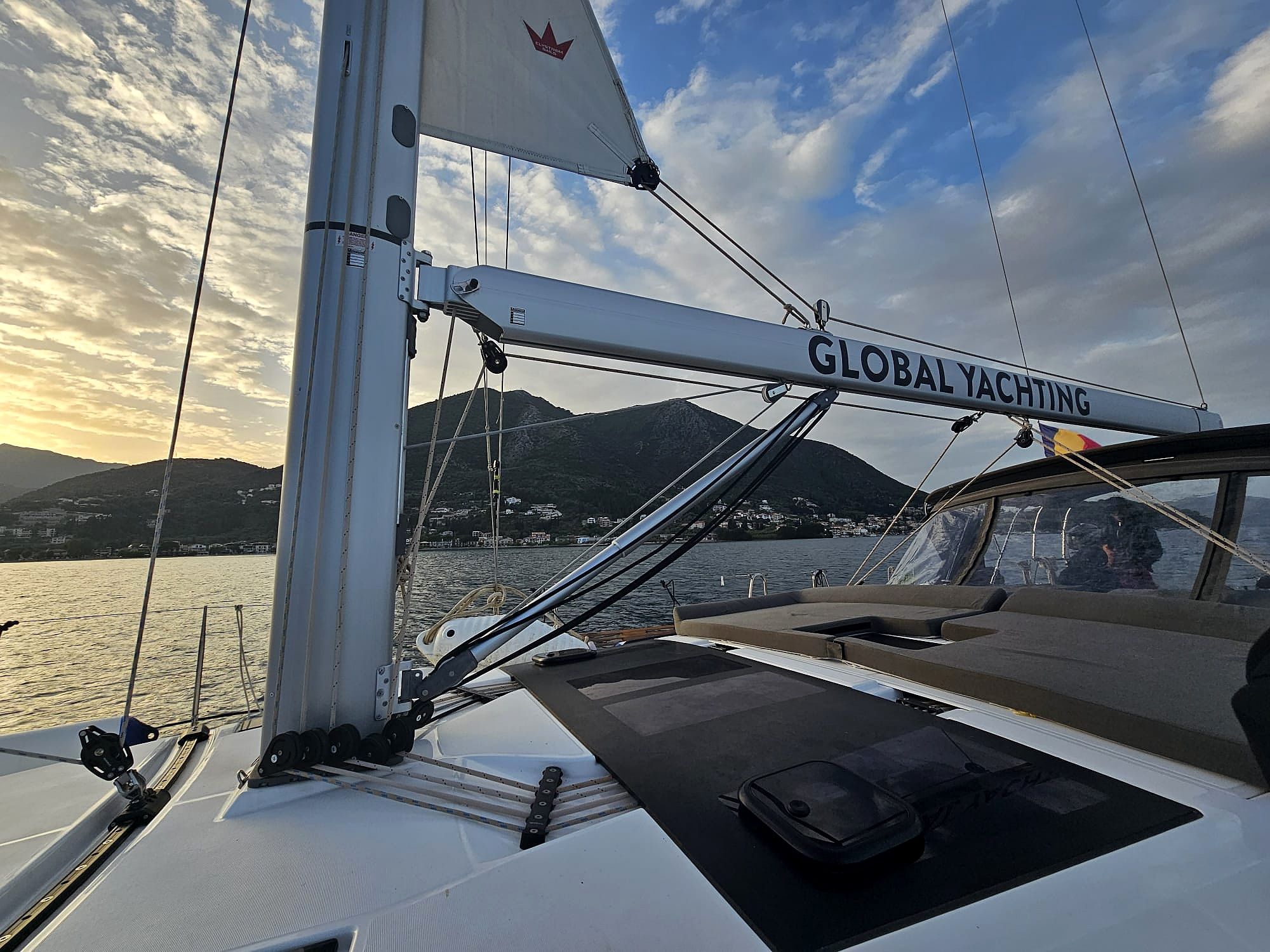 Eine Segelyacht mit einem 'GLOBAL YACHTING'-Schild, einem Mast und Blick auf eine gebirgige Küstenlinie.