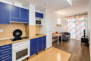 Eine kompakte Küche mit blauen Schränken, einer Mikrowelle, einem Herd und einem Kühlschrank. Es gibt eine Essecke mit Tisch und Stühlen sowie ein Sofa im Wohnzimmer.